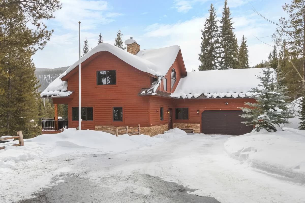 Red wooden house with snow-covered roof surrounded by evergreen trees in winter