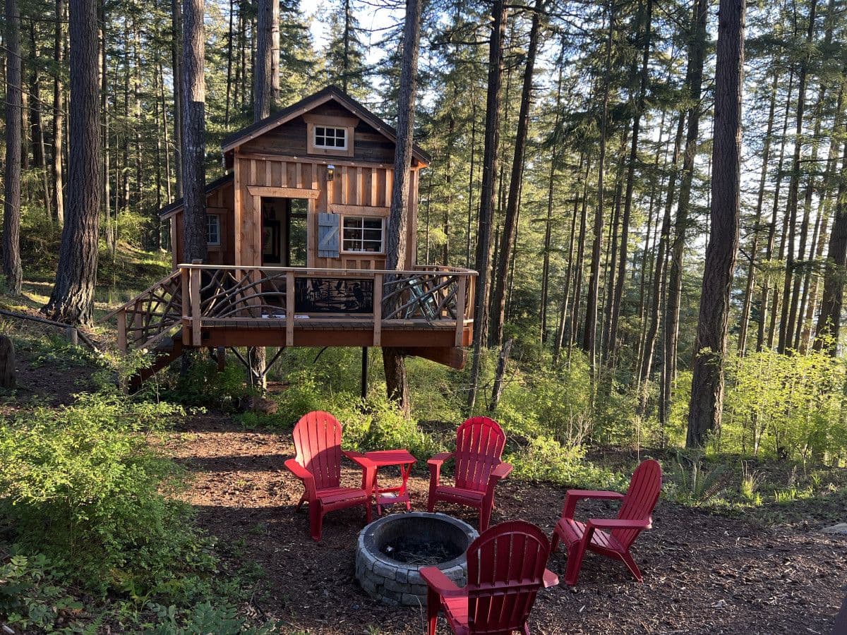 Elevated wooden treehouse with deck surrounded by tall trees and red Adirondack chairs by fire pit