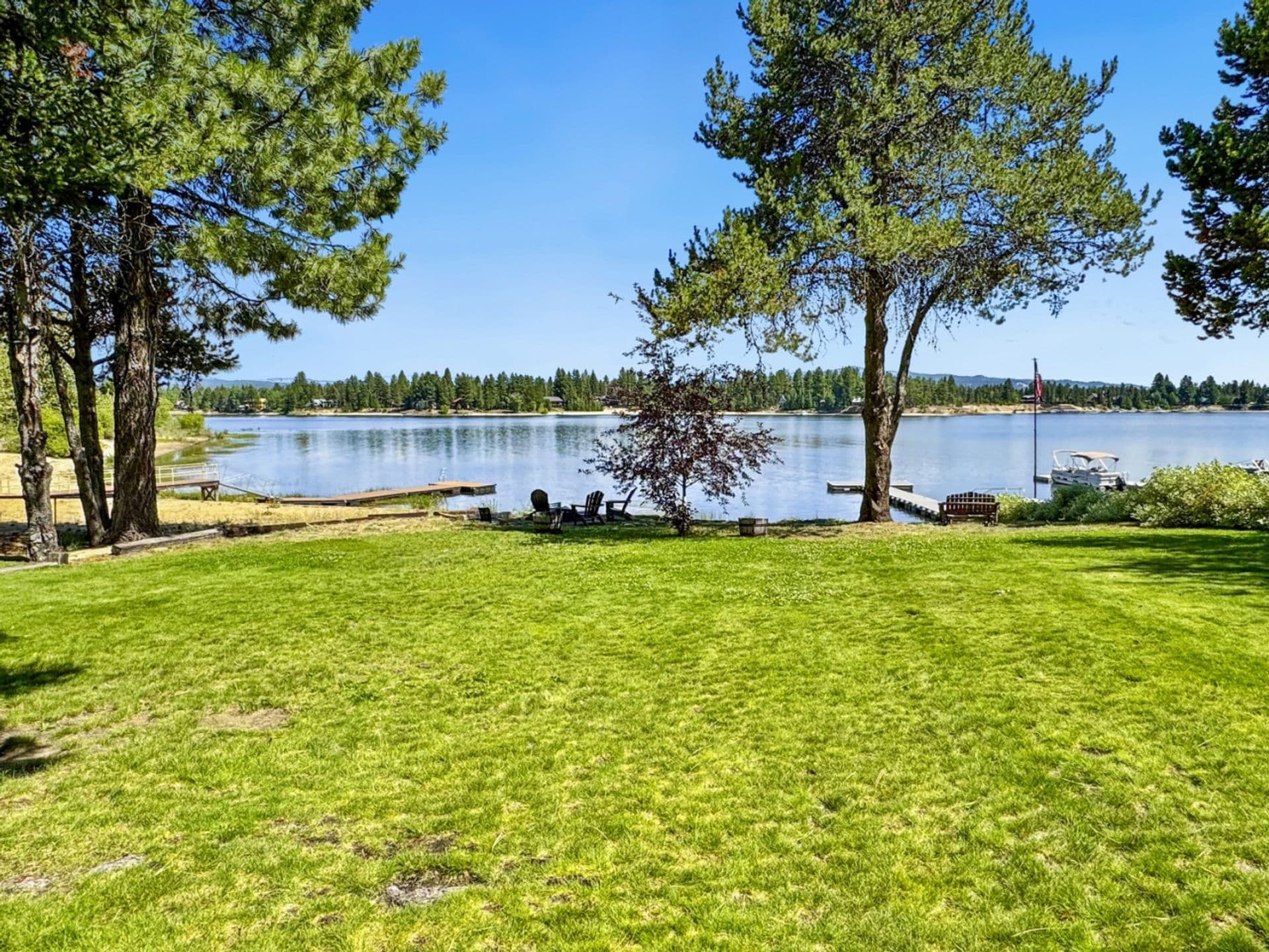 Lakefront lawn with tall pines, boats docked, and distant forested shoreline under clear sky