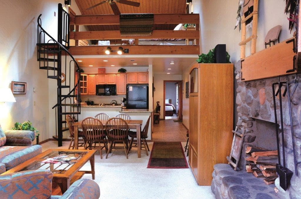 Open-concept lodge interior with wooden beams, spiral staircase, dining area, and kitchen