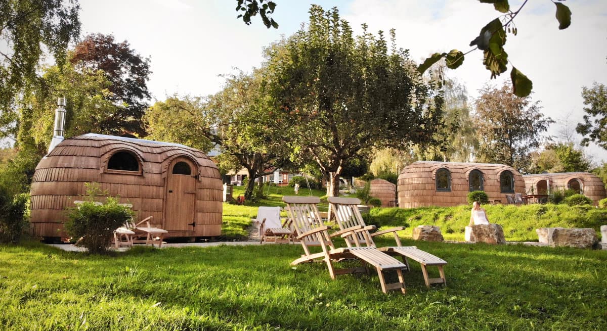 Wooden barrel-shaped glamping pods with lounge chairs on landscaped grounds surrounded by trees