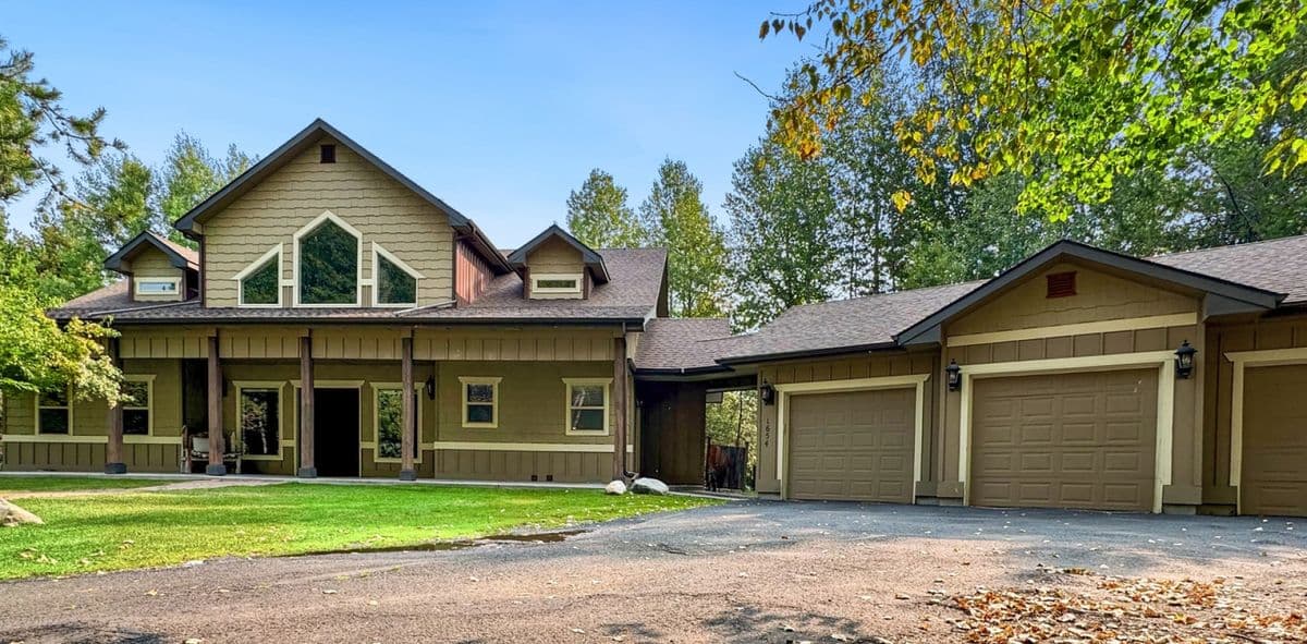 Modern lodge exterior with olive siding, large windows, covered porch, and multiple garages surrounded by pine trees