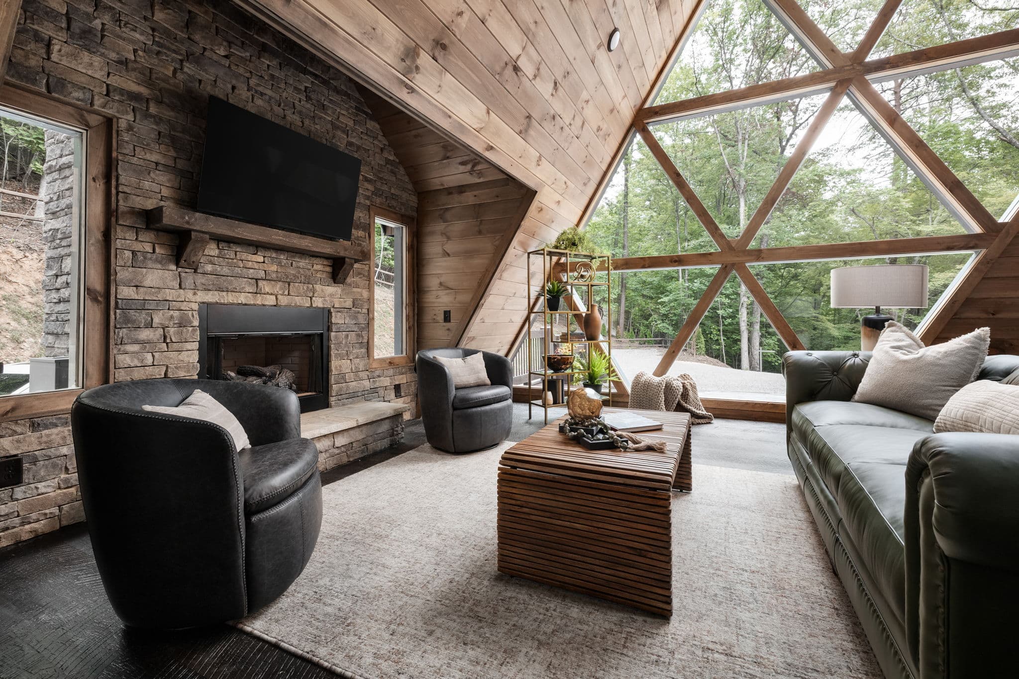Modern living room with fireplace, black leather seating, wood ceiling, and large windows overlooking forest