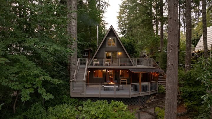 Modern A-frame cabin lit from within, nestled among tall trees with wraparound deck