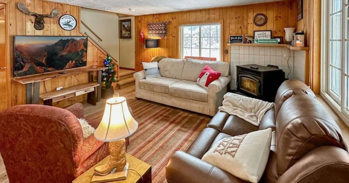 Cozy lodge-style living room with wood paneling, stone fireplace, leather seating, and mountain view on TV