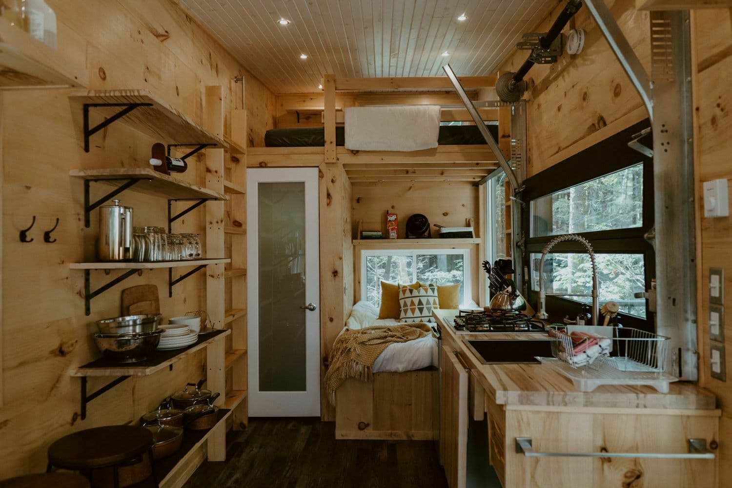 Bright wooden cabin interior with kitchen area, bed with window view, and shelving