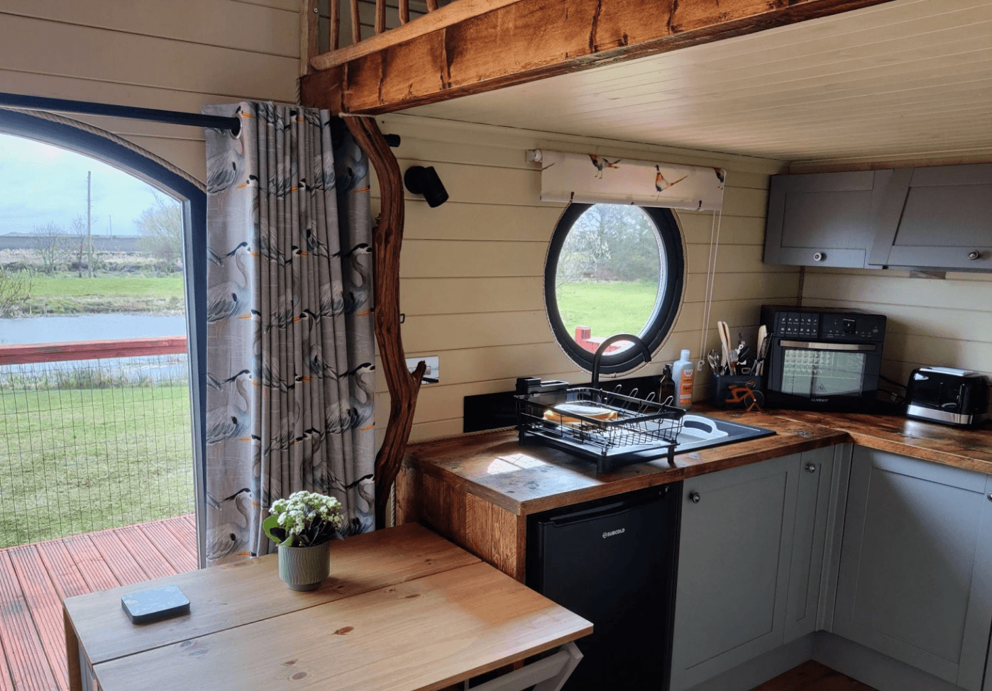 Interior of eco lodge with wooden kitchen, dining table, and large windows overlooking farmland