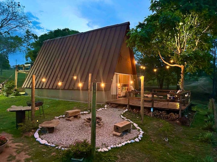 Modern A-frame cabin with metal roof and warm lighting on wooden deck at dusk, surrounded by green lawn and trees