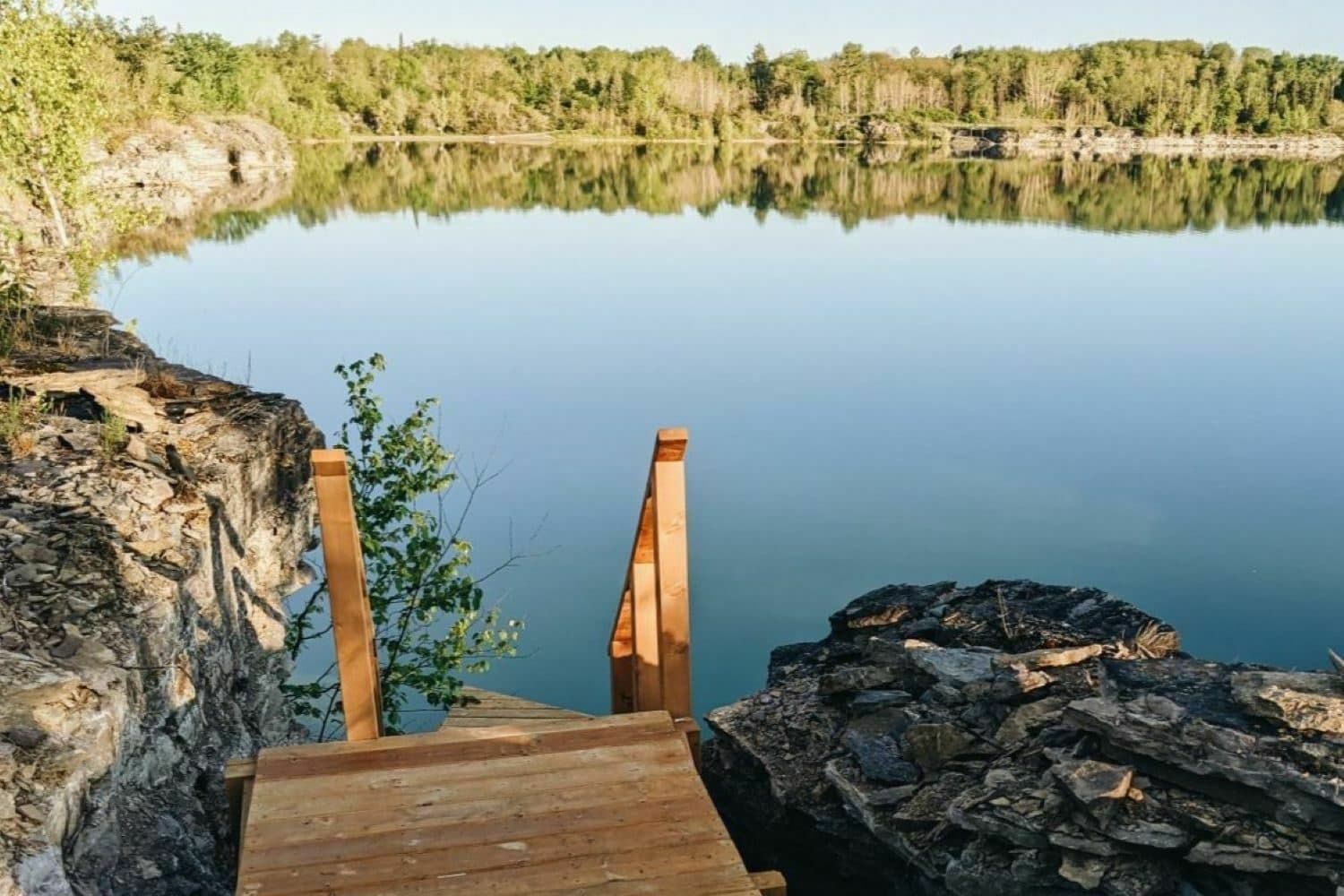 Wooden dock extending into calm quarry lake surrounded by forest and rocky cliffs