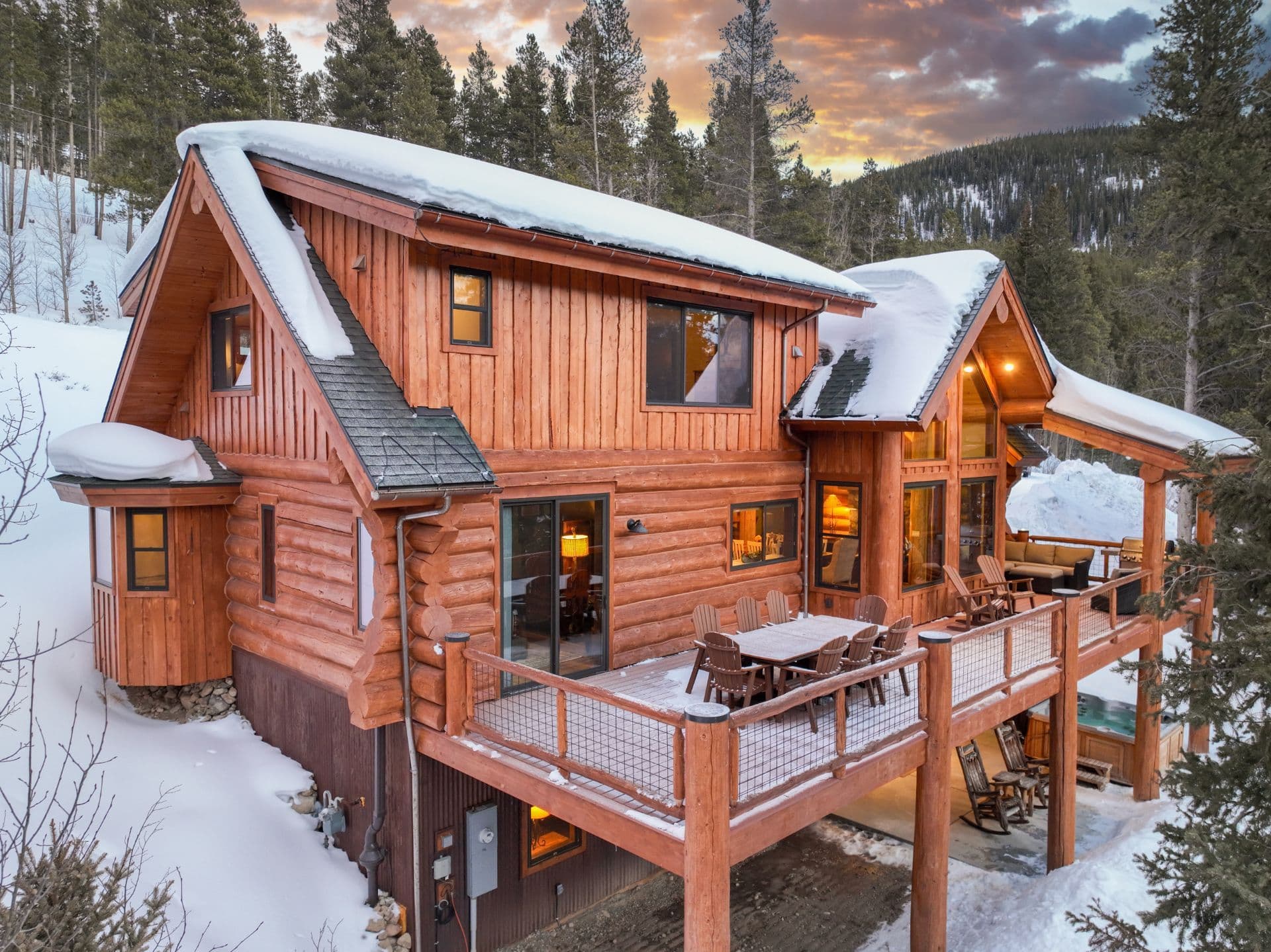 Snow-covered log cabin with large deck and glowing windows in forested mountain setting