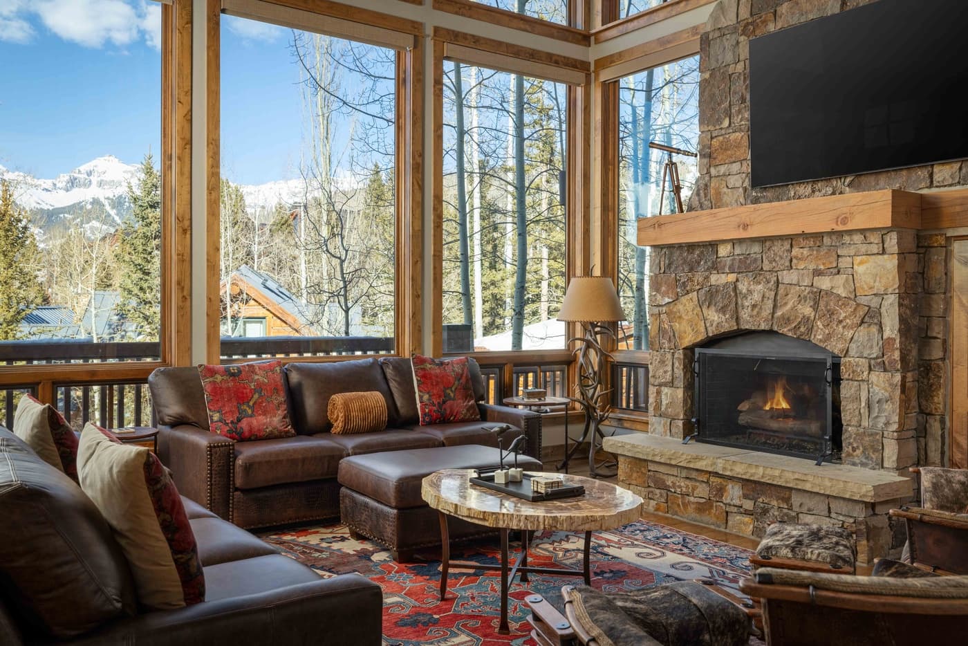 Luxury living room with leather seating, stone fireplace, and floor-to-ceiling windows overlooking snowy mountains