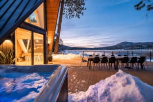 Modern A-frame cabin with illuminated interior, deck seating, and mountain lake view at dusk
