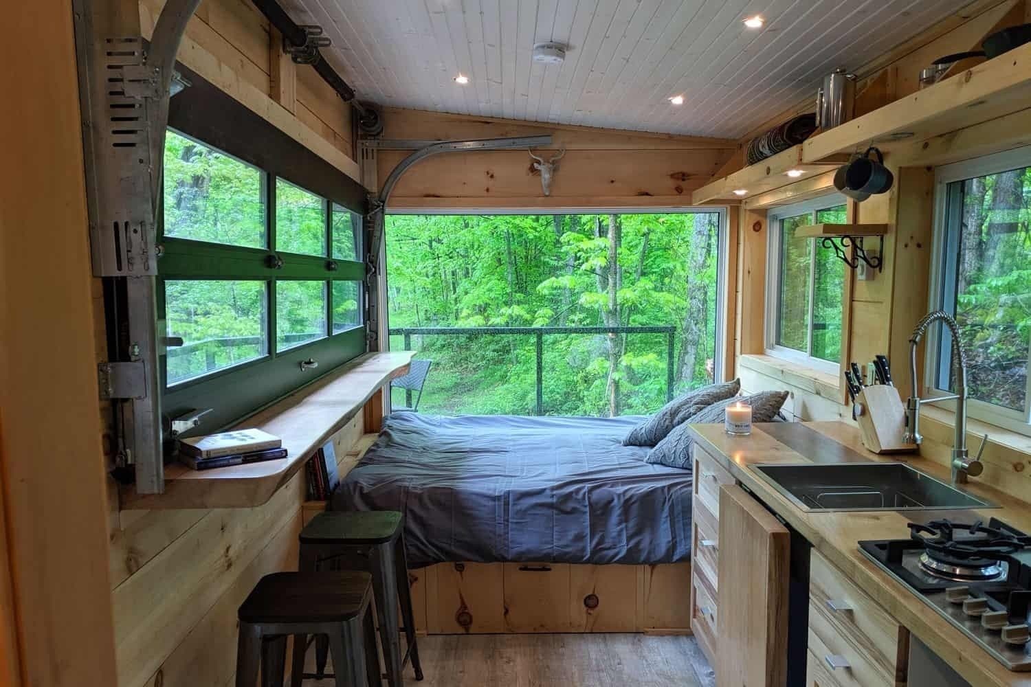 Bright cabin interior with bed, kitchenette, windows overlooking forest and deck
