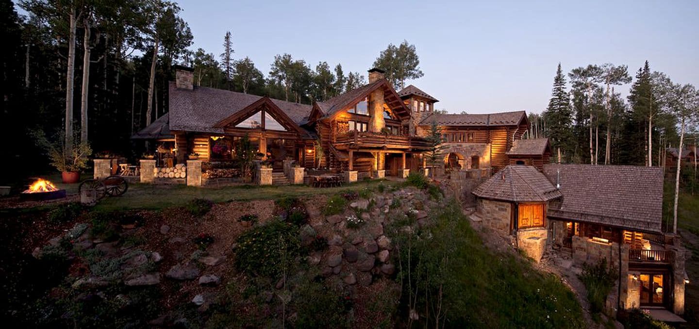Illuminated log cabin resort complex at dusk surrounded by forest and rocky terrain
