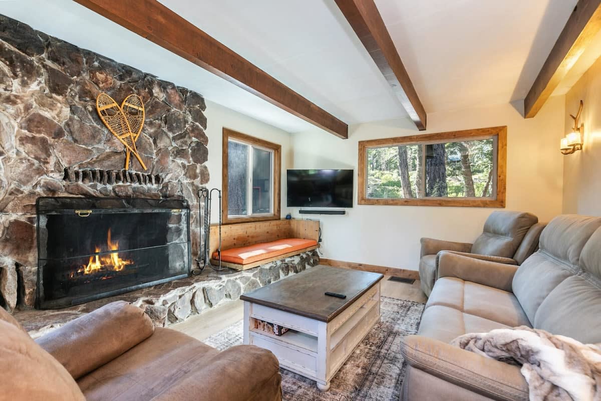 Cozy cabin living room with stone fireplace, wood beams, leather seating, and forest views