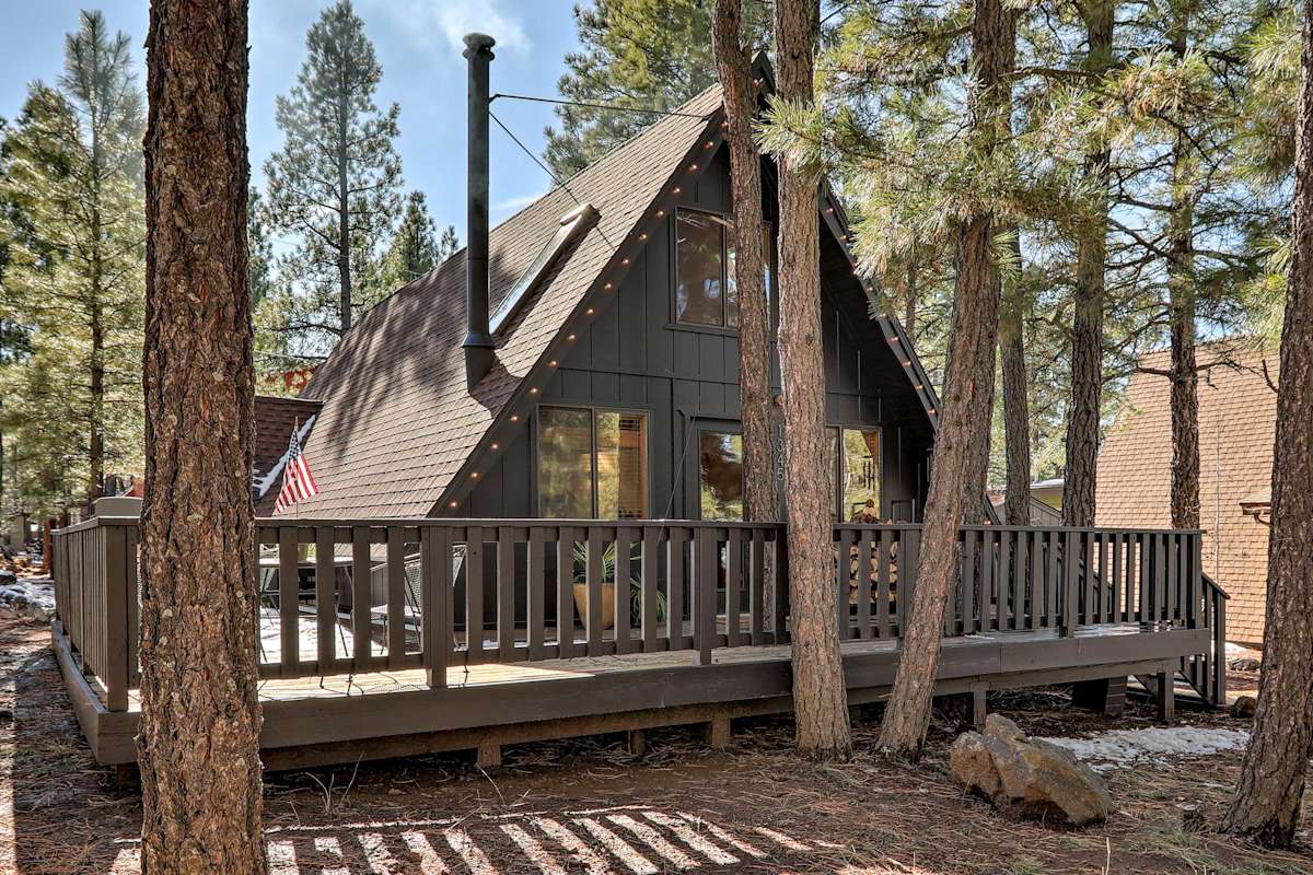 Modern A-frame cabin with dark exterior and wrap-around deck nestled among tall pine trees