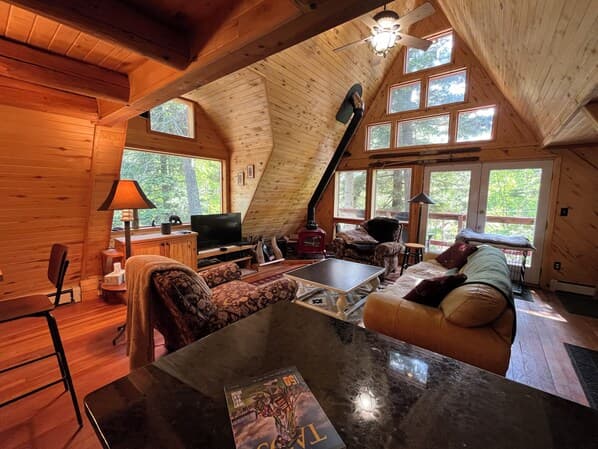 Bright wood-beamed A-frame cabin interior with large windows overlooking forest, furnished with lounge areas and fireplace.
