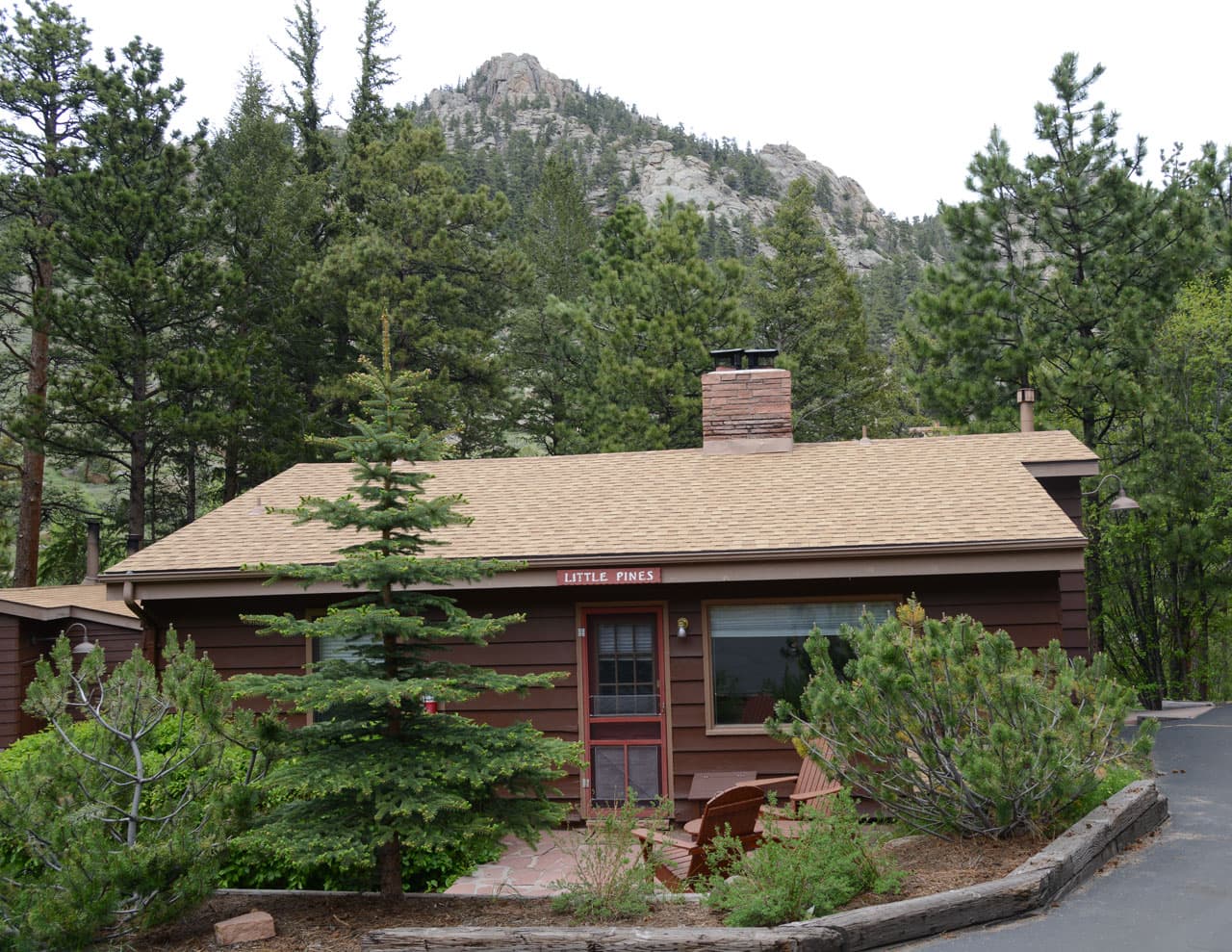 Brown lodge building with red door and chimney set among pine trees with rocky mountain backdrop