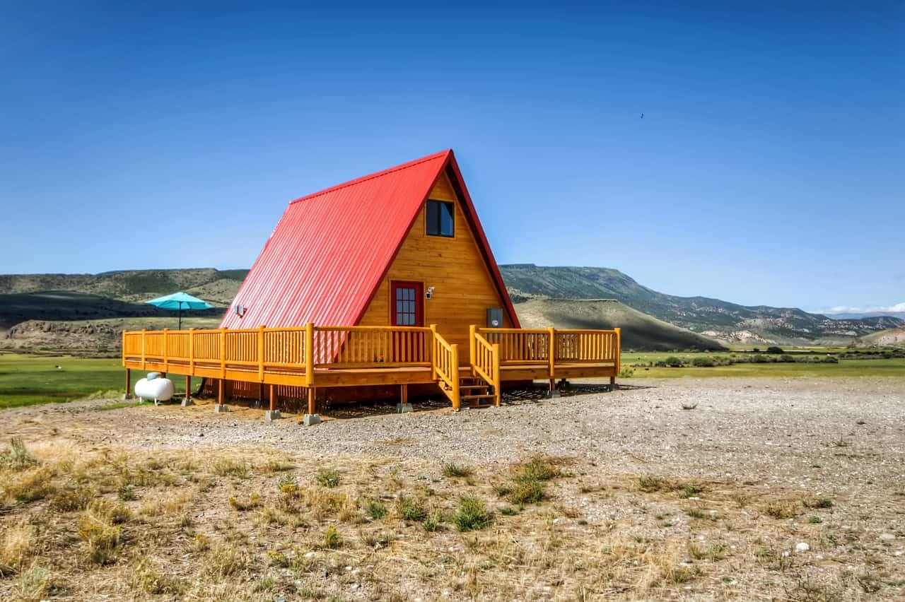 Wooden A-frame cabin with red roof and yellow deck in mountain valley landscape