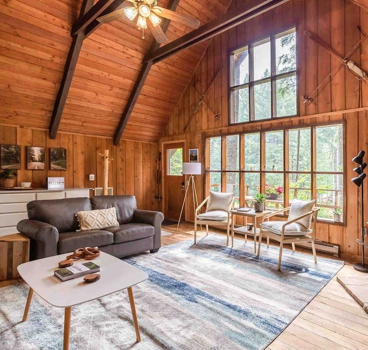 Wood-paneled A-frame interior with leather sofa, large windows, and modern furnishings