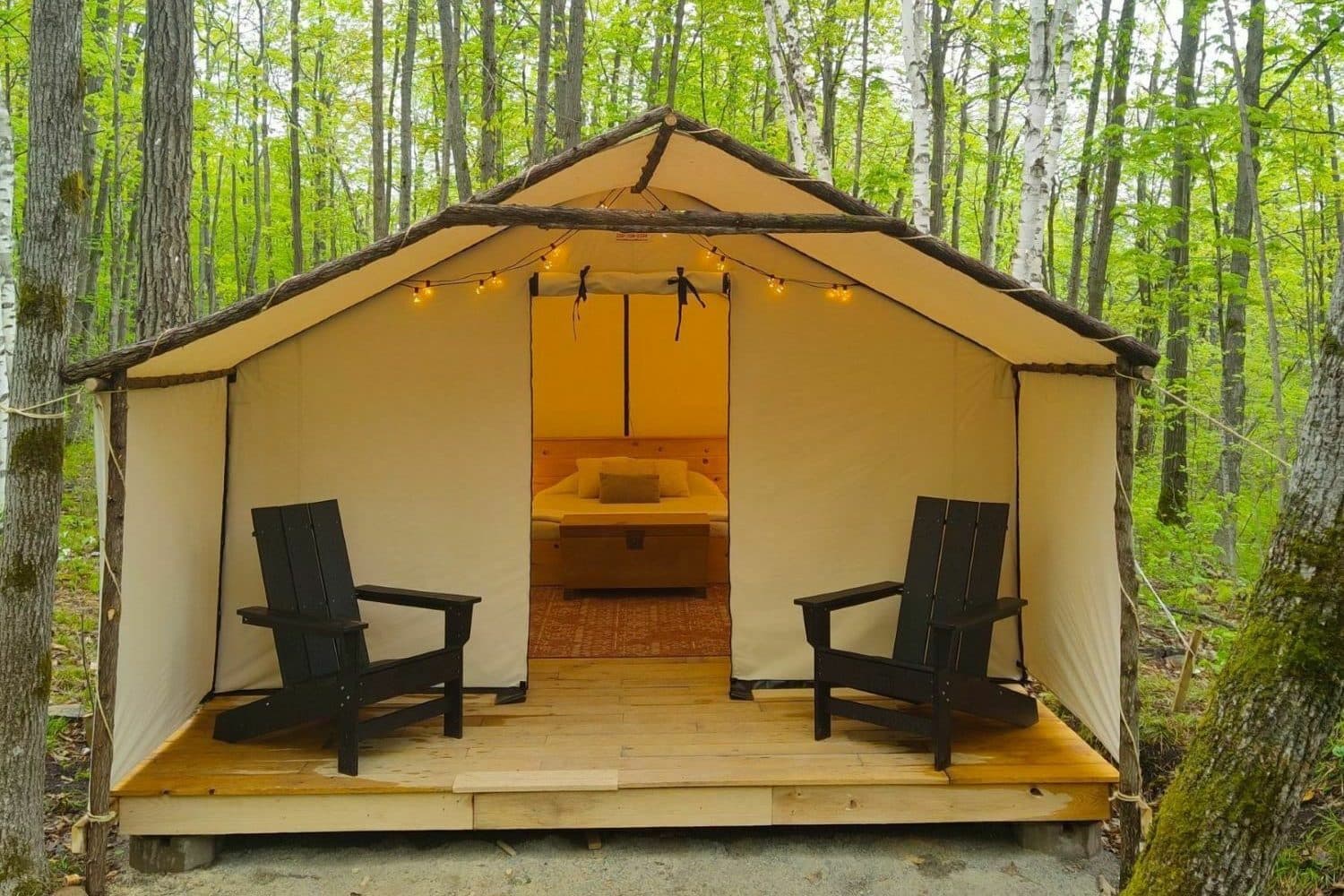 Glamping tent with canvas walls, wooden deck, two black chairs, and bed visible inside forest setting