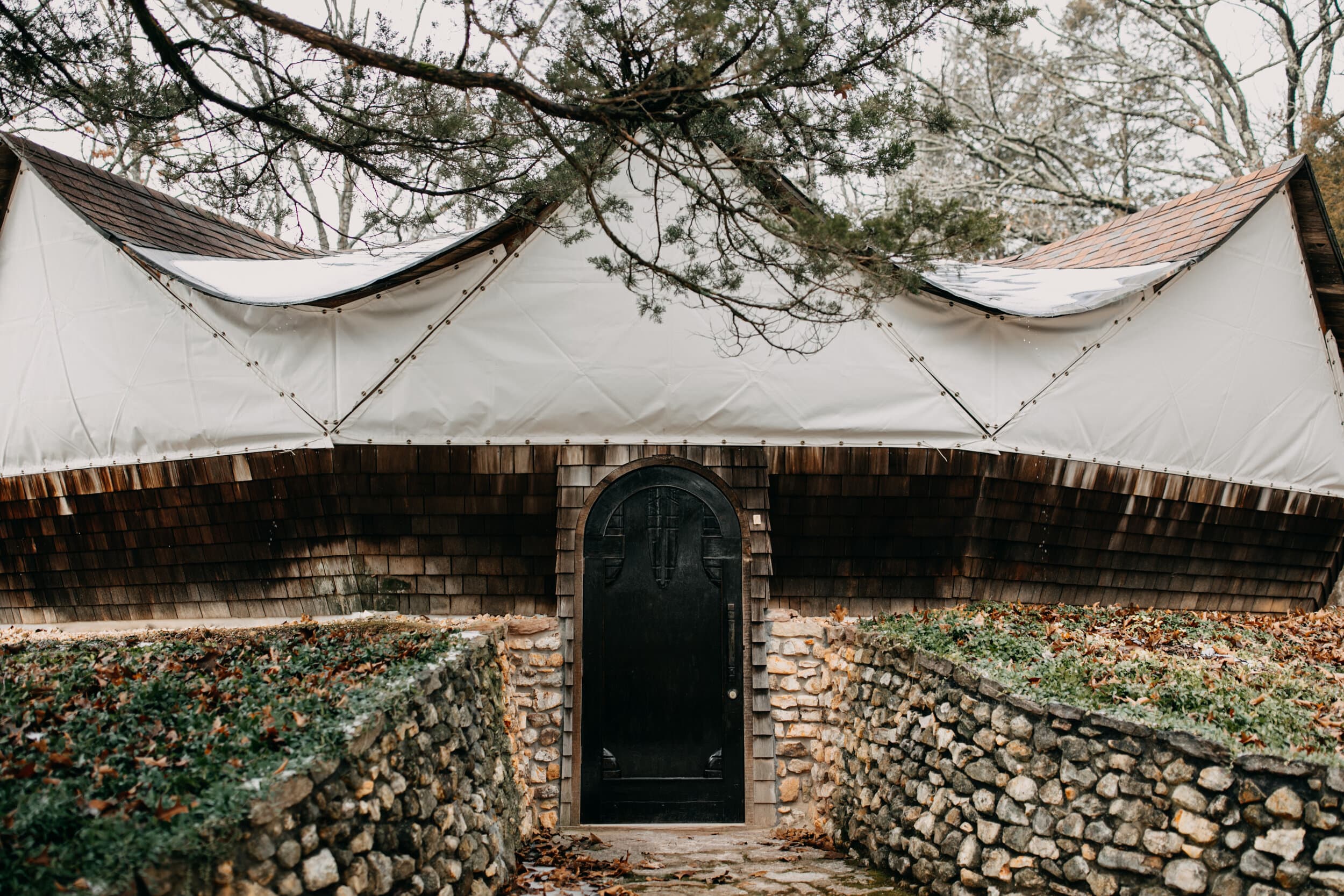 Stone yurt structure with black door, white canvas roof, and bare tree branches overhead