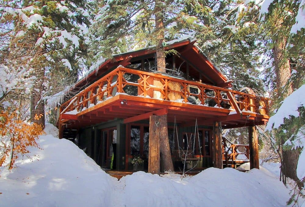 Rustic wooden treehouse with red-orange railings nestled among tall pine trees in winter snow