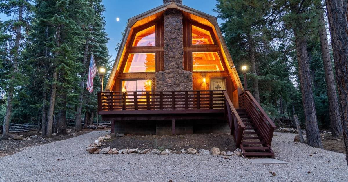 A-frame cabin at dusk with warm interior lighting, stone chimney, and wooden deck surrounded by tall pine trees