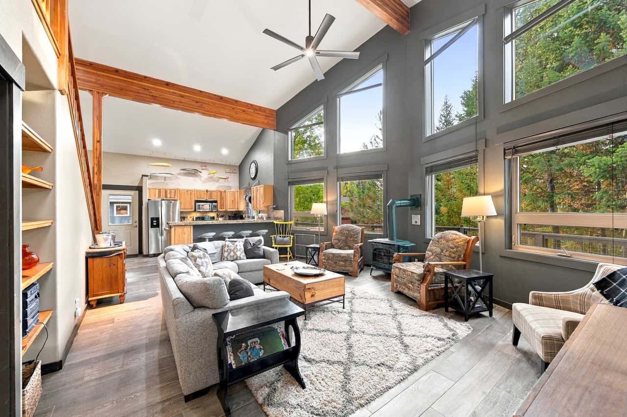 Open-plan living room and kitchen with high ceilings, wood beams, large windows overlooking forest, and modern furnishings