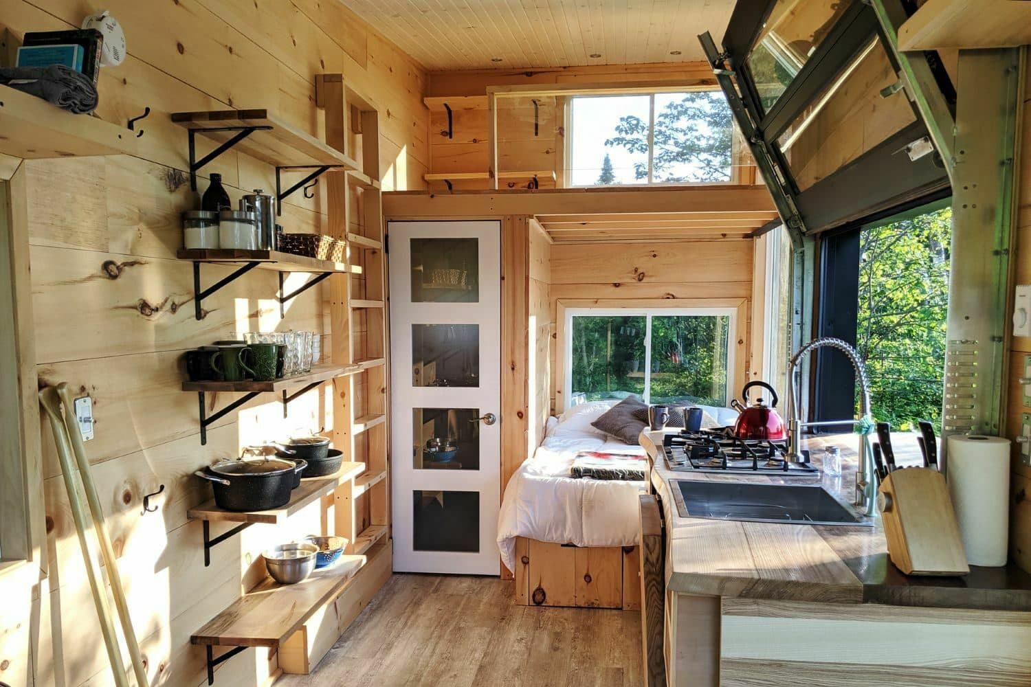 Interior of a modern wooden cabin with built-in kitchen, bed, and large windows overlooking forest