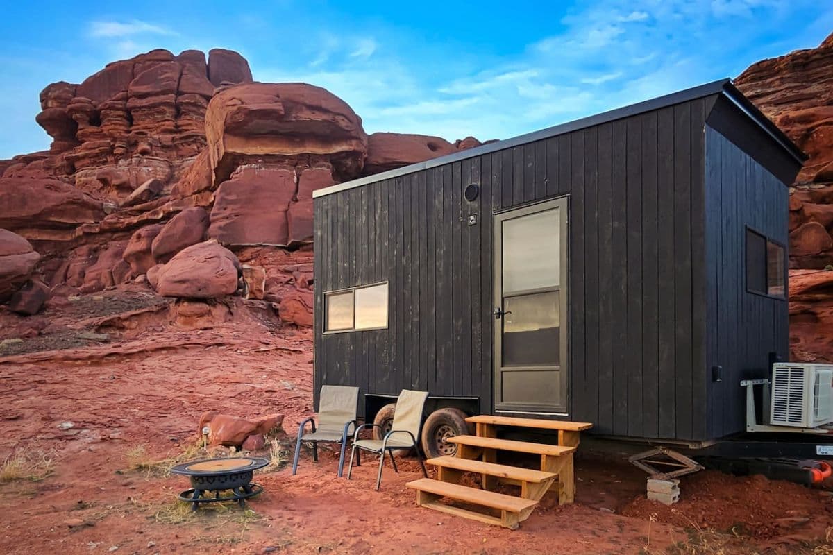 Black cabin with wooden steps nestled against dramatic red rock formations under blue sky