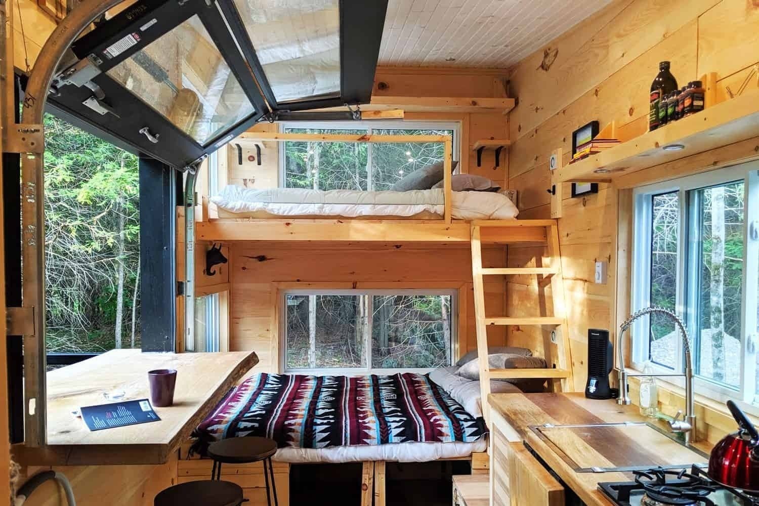 Interior of a wooden cabin with bunk beds, desk, kitchenette, and forest views through windows