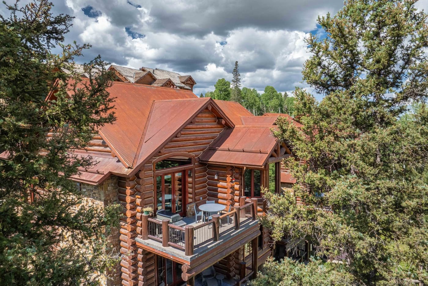 Rustic log lodge with copper metal roof nestled among pine trees under cloudy sky