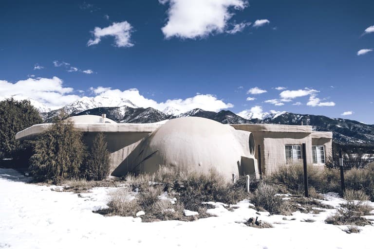 Curved dome structure with snow and mountains in background under blue sky