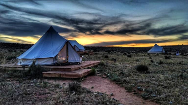Blue bell tent glowing at dusk with wooden platform and desert landscape at sunset