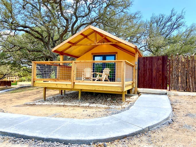 Yellow glamping cabin with covered porch and Adirondack chairs on concrete pad
